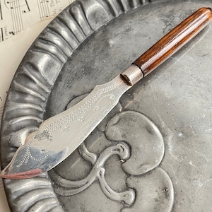 May include: A vintage butter knife with a silver blade and a dark wood handle. The blade features an etched design. The knife rests on an antique metal plate. The handle is approximately 7.6 cm long.