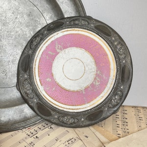 May include: A round metal tray featuring a dark, tarnished silver-toned border with embossed floral motifs and a central circular inlay. The inlay displays a white centre surrounded by a pink band with a gold-toned decorative pattern and trim.