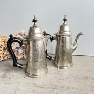 Könnte beinhalten: Zwei silberne Teekannen mit schwarzen Griffen und verzierten Blumendesigns. Die Teekannen haben eine polierte Oberfläche und sind in ausgezeichnetem Zustand.