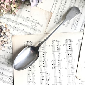 Op de afbeelding: Een lange, zilveren lepel met een afgeronde kom en een slanke steel. De lepel ligt op een verzameling vintage bladmuziek met bloemaccenten. De lepel heeft gegraveerde initialen.