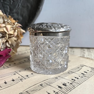 May include: A clear, cut-glass jar with a silver lid. The jar has a geometric pattern and a decorative silver lid with an embossed floral design. The jar is sitting on a sheet of vintage sheet music.