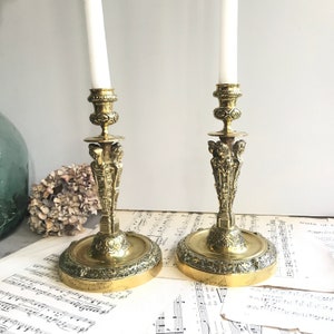 May include: Two matching brass candlesticks with white taper candles. Each candlestick features a circular base with ornate embossed detailing, a central stem adorned with sculpted figures, and a decorative top cup for holding the candle securely.