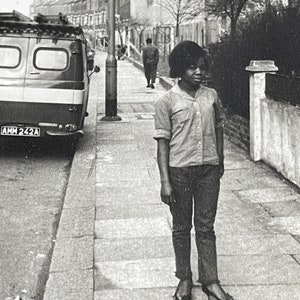 May include: Black and white photograph of a person standing on a sidewalk. The person is wearing a button-down shirt, dark pants, and heels. A van and buildings are in the background. The image is taken outdoors on a cloudy day.