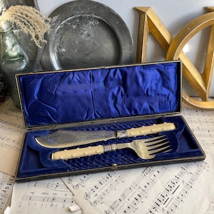 May include: A vintage silver knife and fork set with ivory handles, displayed in a blue velvet lined box. The box is made of black leather and has a gold trim.