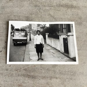 May include: Black and white photograph of a person standing on a sidewalk next to a street with a van parked on the side. The person is wearing a light-colored button-down shirt and a dark skirt. The image is a vintage snapshot.