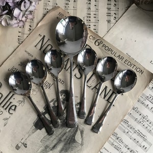 May include: A collection of vintage silver spoons arranged on a backdrop of aged sheet music and a newspaper clipping. The spoons vary in size, with ornate detailing on the handles. The reflective surfaces of the spoons mirror the surrounding environment.