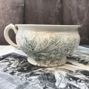 May include: Vintage white ceramic chamber pot with a handle and a green floral pattern. The pot has a wide opening and a small pedestal base. The floral design is detailed and covers the exterior. A classic piece of antique homeware.