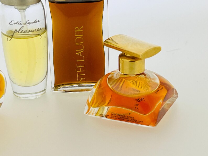 May include: Three bottles of perfume. The bottle on the left is clear glass with a gold cap and the text "Est&eacute;e Lauder pleasures" printed on the label. The bottle in the centre is clear glass with a gold cap and the text "ESTEE LAUDER" printed on the label. The bottle on the right is clear glass with a gold cap and a gold top. The bottle is filled with a light amber liquid.