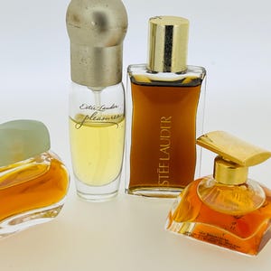 May include: Four bottles of perfume, two with gold caps and two with silver caps. The bottles are filled with a light yellow liquid. The bottles are labelled "Estee Lauder" and "Pleasures".