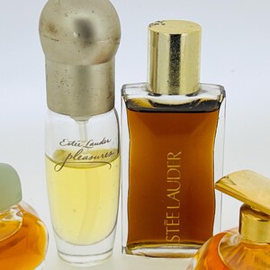 May include: Three glass bottles of perfume. The bottle on the left is clear with a gold cap and the text "Est&eacute;e Lauder pleasures" on the label. The bottle in the centre is clear with a gold cap and the text "ESTEE LAUDER" on the label. The bottle on the right is clear with a gold cap.