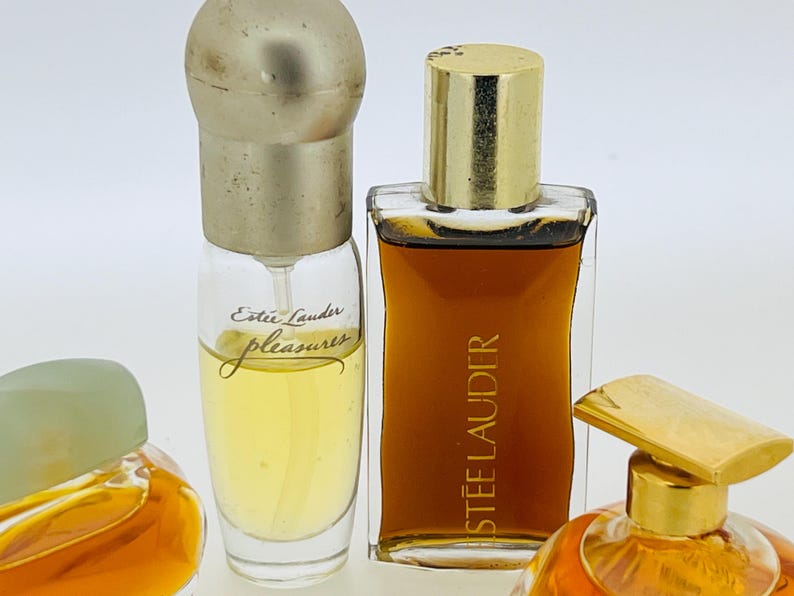 May include: Three glass bottles of perfume. The bottle on the left is clear with a gold cap and the text "Est&eacute;e Lauder pleasures" on the label. The bottle in the centre is clear with a gold cap and the text "ESTEE LAUDER" on the label. The bottle on the right is clear with a gold cap and a gold top.