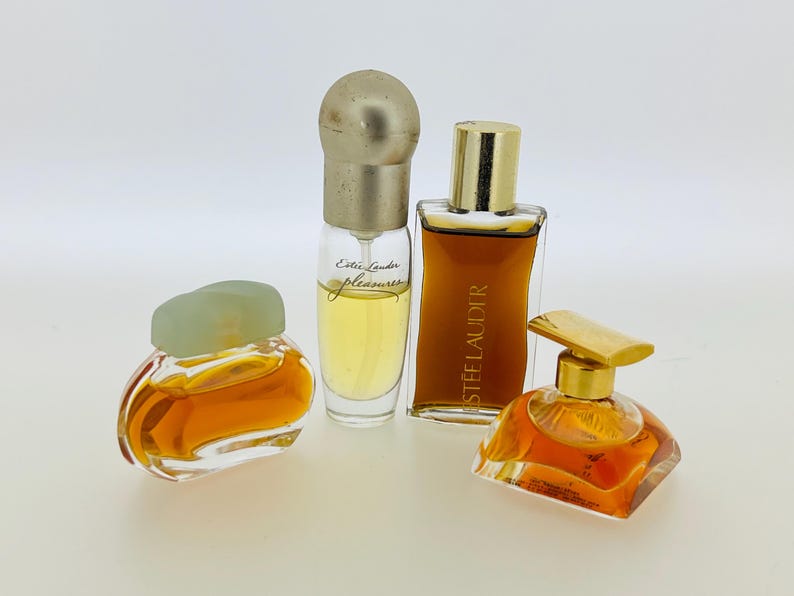May include: Four bottles of perfume, two with gold caps, one with a green cap, and one with a clear cap. The bottles are filled with a light amber liquid. The bottles are labelled "Estee Lauder" and "Est&eacute;e Lauder Pleasures".