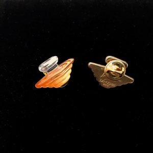 May include: A gold-coloured pin featuring a clear and orange glass perfume bottle design. The reverse of the pin reads "Lancôme Paris".