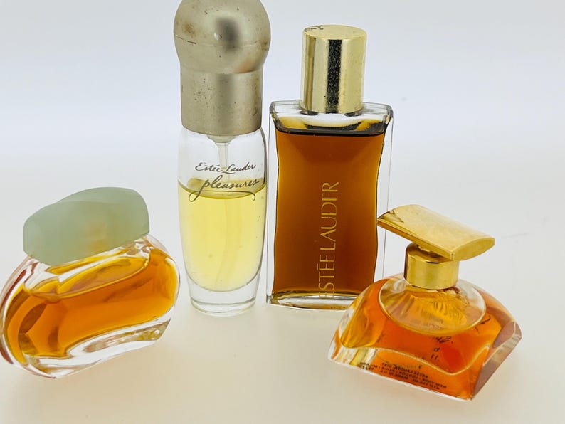 May include: Four bottles of perfume, two clear glass bottles with gold caps, one clear glass bottle with a green cap, and one amber glass bottle with a gold cap. The bottles are all filled with liquid and have labels with the name "Estee Lauder" on them.
