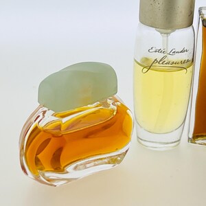May include: Three miniature bottles of perfume. The bottle on the left is clear glass with a light green cap and a gold liquid inside. The middle bottle is clear glass with a silver cap and a yellow liquid inside. The bottle on the right is clear glass with a silver cap and a brown liquid inside. The middle bottle has the text "Est&eacute;e Lauder pleasures" printed on the label.