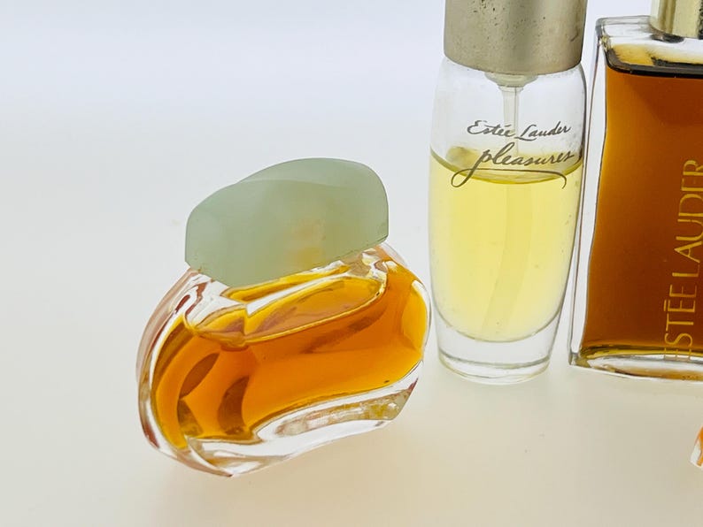 May include: Three miniature bottles of perfume. The bottle on the left is clear glass with a light green cap and a gold liquid inside. The middle bottle is clear glass with a silver cap and a yellow liquid inside. The bottle on the right is clear glass with a silver cap and a brown liquid inside. The middle bottle has the text "Est&eacute;e Lauder pleasures" printed on the label.