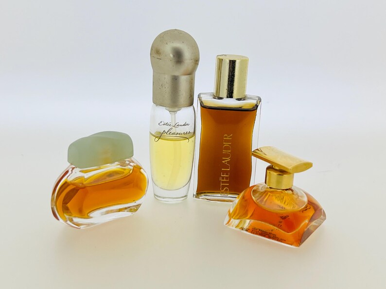 May include: Four bottles of perfume, two with clear glass and gold caps, one with a green cap and clear glass, and one with a gold cap and clear glass. The bottles are filled with a light amber liquid. The bottles are labelled "Estee Lauder Pleasures" and "Estee Lauder".