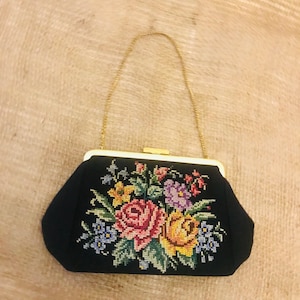 Könnte beinhalten: Schwarze Clutch-Tasche mit goldener Kette und Verschluss. Die Tasche hat ein farbenfrohes Blumen-Kreuzstich-Design mit rosa, gelben, lila und blauen Blumen. Der Rahmen ist weiß.