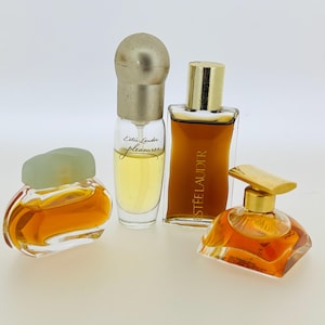 May include: Four bottles of perfume, two with clear glass and gold caps, one with a green cap and clear glass, and one with a gold cap and clear glass. The bottles are filled with a light amber liquid. The bottles are labelled "Estee Lauder Pleasures" and "Estee Lauder".