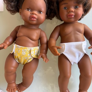 May include: Two dark-skinned baby dolls with brown hair and realistic features. One doll wears a yellow floral diaper, the other a white diaper. The dolls are posed side-by-side, showcasing their detailed facial expressions and body shapes.