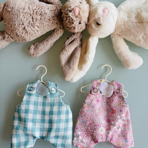 May include: Two plush bunny rabbits with long ears are positioned above two baby outfits. One outfit is blue and white checkered, the other is pink with a floral pattern. Both outfits are on white hangers.