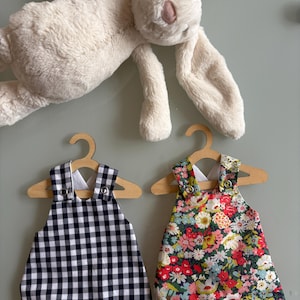 May include: A cream-coloured plush rabbit toy positioned above two small rompers hanging on wooden hangers. One romper features a black and white gingham check pattern, while the other displays a vibrant floral print with red, pink, and green flowers.
