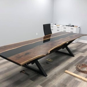 Op de afbeelding: Een lange, donkere houten tafel met een zwarte epoxyrivier die door het midden loopt. De tafel heeft een levende rand en wordt ondersteund door een zwart metalen X-vormig onderstel.