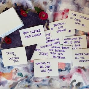 Peut inclure: Un jeu de cartes rectangulaires blanches avec du texte manuscrit violet, incluant des phrases comme 'Your silence says enough' et 'Beautiful and vibrant', disposées autour d'une pochette en tissu violet foncé avec deux petites fleurs rouges.