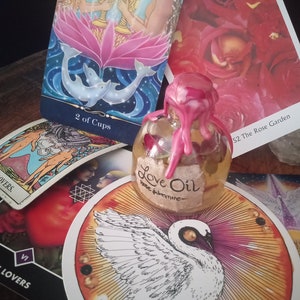 May include: Tarot cards with a pink and blue image of two people holding cups, a white swan card, and a small bottle of love oil with a pink wax seal. The bottle label reads "Love Oil Rose Jasmine".