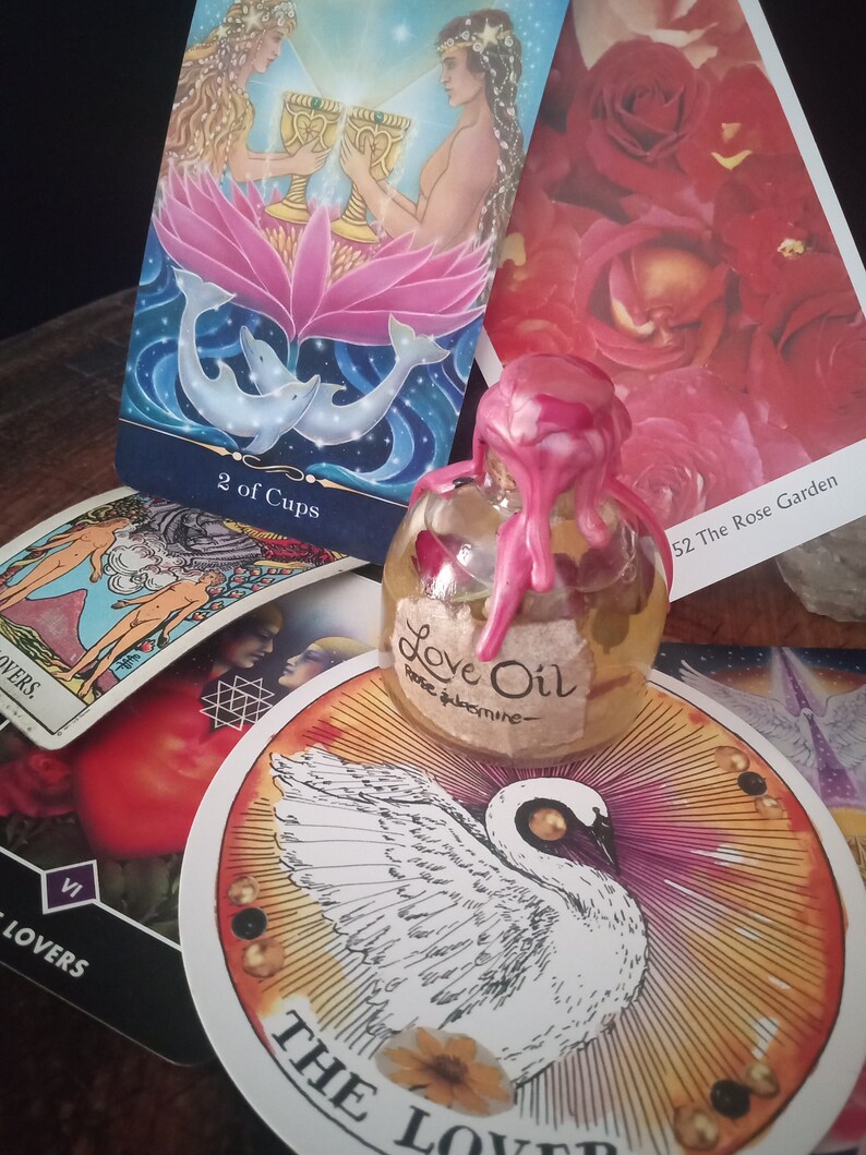 May include: Tarot cards with a pink and blue image of two people holding cups, a white swan card, and a small bottle of love oil with a pink wax seal. The bottle label reads "Love Oil Rose Jasmine".
