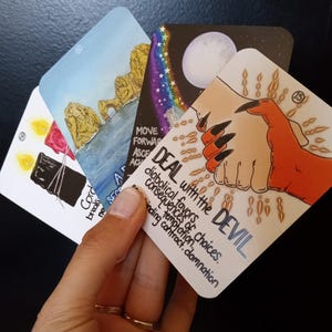 May include: A collection of tarot cards with various illustrations and text. One card features a hand reaching out to another with the words "DEAL with the DEVIL." Other cards show images of a seascape, a night sky, and candles.
