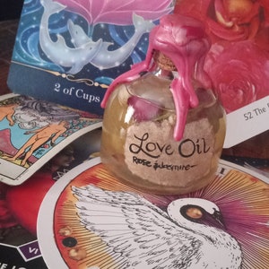 May include: A clear glass bottle with a pink wax seal and a label that reads "Love Oil Rose & Jasmine". The bottle is sitting on a white and yellow circular card with a black and white illustration of a swan.
