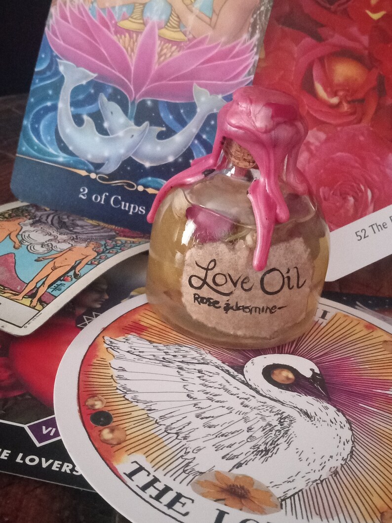 May include: A clear glass bottle with a pink wax seal and a label that reads "Love Oil Rose & Jasmine". The bottle is sitting on a white and yellow circular card with a black and white illustration of a swan.