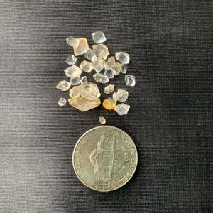 May include: A small pile of clear and light amber colored crystals, some with a slight yellow tint, are scattered on a black surface. A silver US quarter is below the crystals for size comparison.