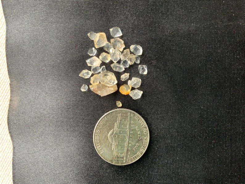 May include: A small pile of clear and light amber colored crystals, some with a slight yellow tint, are scattered on a black surface. A silver US quarter is below the crystals for size comparison.