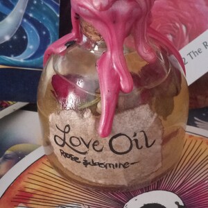 May include: A glass bottle with a pink wax seal, filled with a clear liquid and containing dried flowers. The bottle has a label that reads "Love Oil Rose & Jasmine".