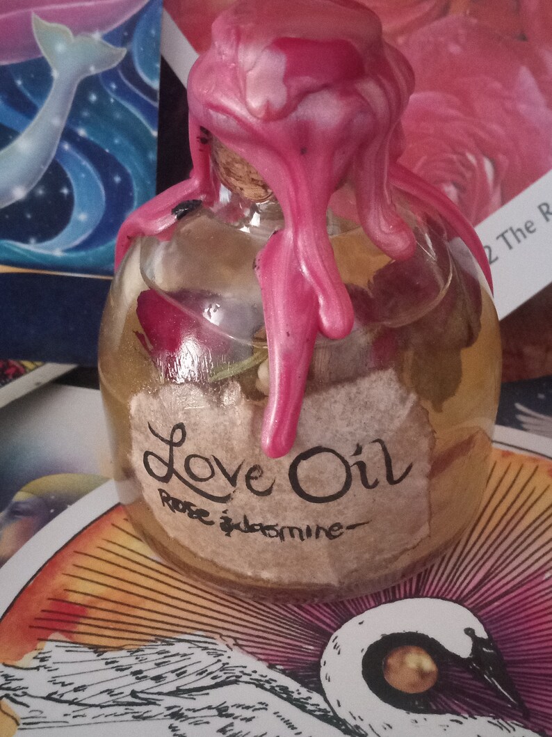 May include: A glass bottle with a pink wax seal, filled with a clear liquid and containing dried flowers. The bottle has a label that reads "Love Oil Rose & Jasmine".