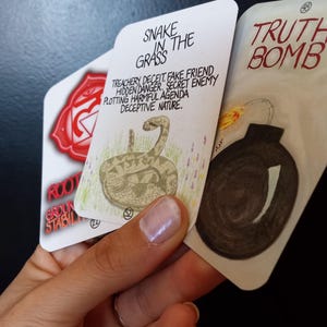 Puede incluir: Una mano sosteniendo tres cartas de tarot. La carta superior dice "SNAKE IN THE GRASS" con una ilustración de serpiente y texto sobre engaño. La segunda carta tiene un símbolo de chakra rojo. La tercera carta dice "TRUTH BOMB" con una ilustración de bomba.