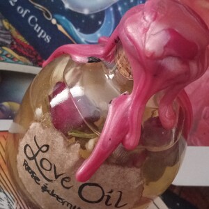 May include: A clear glass bottle with a pink wax seal, filled with a light yellow liquid and a label that reads "Love Oil Rose & Jasmine".