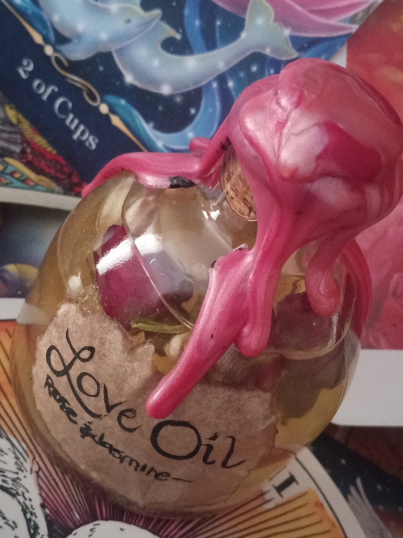 May include: A clear glass bottle with a pink wax seal, filled with a light yellow liquid and a label that reads "Love Oil Rose & Jasmine".