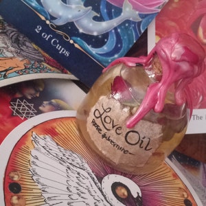 May include: A clear glass bottle with a pink wax seal and a label that reads "Love Oil - Rose & Jasmine". The bottle is sitting on a tarot card with a white bird design.