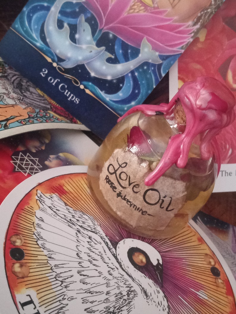 May include: A clear glass bottle with a pink wax seal and a label that reads "Love Oil - Rose & Jasmine". The bottle is sitting on a tarot card with a white bird design.