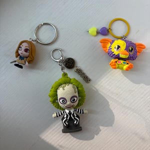 May include: Three character keychains: one with a blonde figure in a tan dress, one with a green-haired figure in a black and white striped suit, and one with a purple and orange creature featuring a yellow ring and heart-shaped beads.