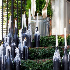 May include: A set of black wine bottles with white candles on a set of brick steps covered in green ivy. The candles are lit and dripping wax.