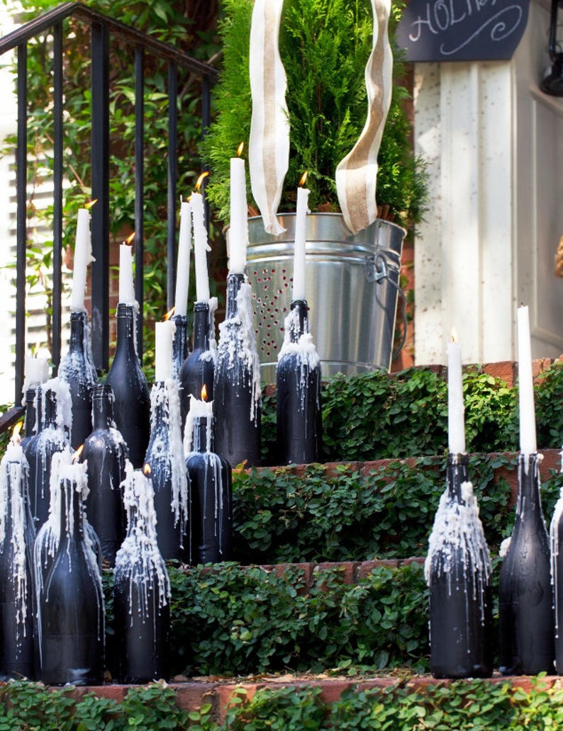 May include: A set of black wine bottles with white candles on a set of brick steps covered in green ivy. The candles are lit and dripping wax.