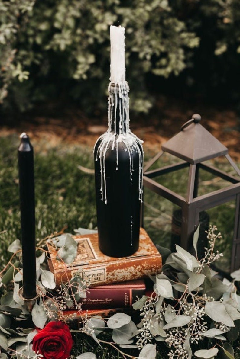 May include: A black glass bottle with a white candle inside, dripping wax. The bottle is sitting on a stack of old books with the text "Express 1897" visible. The bottle is surrounded by greenery.
