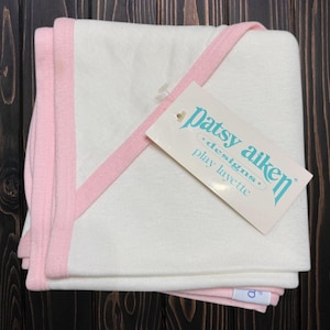 May include: A folded white baby blanket with a pink trim and a tag that reads "patsy aiken designs play layette". The blanket is made of soft material and is folded neatly. The tag is attached with a clear plastic fastener.