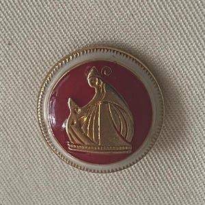 Puede incluir: Un pin redondo con un centro rojo que presenta una ilustración dorada de dos figuras. El pin tiene un borde blanco y un borde dorado. El fondo es de color beige claro.