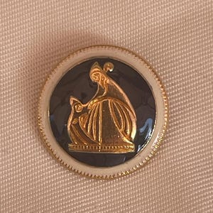 Puede incluir: Un botón redondo con un relieve dorado de dos figuras sobre un fondo azul oscuro, enmarcado por un borde blanco y un borde dorado. El botón está sobre una tela beige claro.