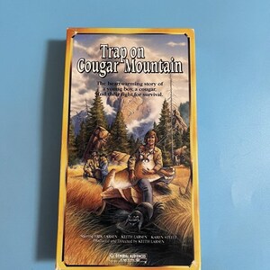Puede incluir: Una cinta VHS titulada "Trap on Cougar Mountain". La portada presenta un puma y un joven en un entorno montañoso. El texto dice: "La conmovedora historia de un niño, un puma y su lucha por la supervivencia."